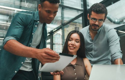 Searching for solutions. Group of three young and positive employees using modern technologies and discussing something while working in modern office. Job concept. Workplace